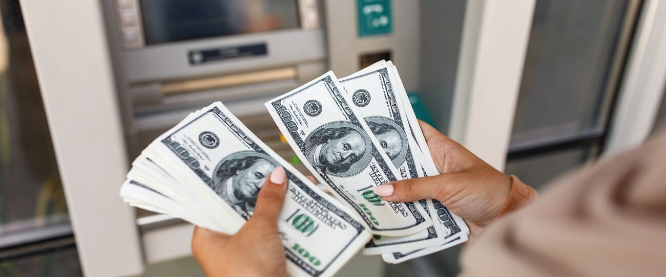 A pair of hands holding a fan of banknotes in front of an ATM.