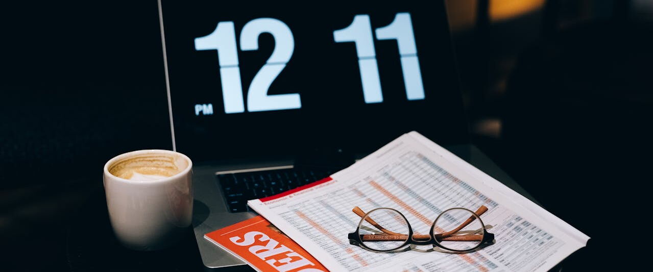 A desk with an open laptop showing a large flip clock, a cup of coffee, reading glasses and a printed spreadsheet.