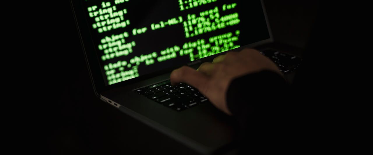 A hand resting on a laptop keyboard in the dark, the screen filled with green code.