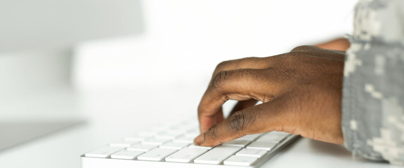 A hand in a camouflage uniform sleeve typing on a white keyboard beside a monitor.