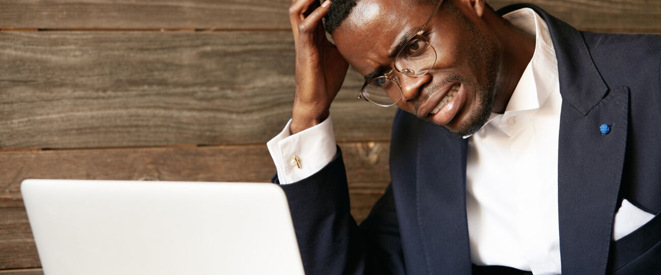 A man in a suit and glasses sits at a laptop with a hand to his head, looking dismayed.
