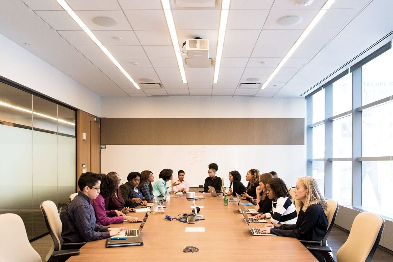 Directors seated along a boardroom table with laptops open, listening to a colleague at the head of the room.