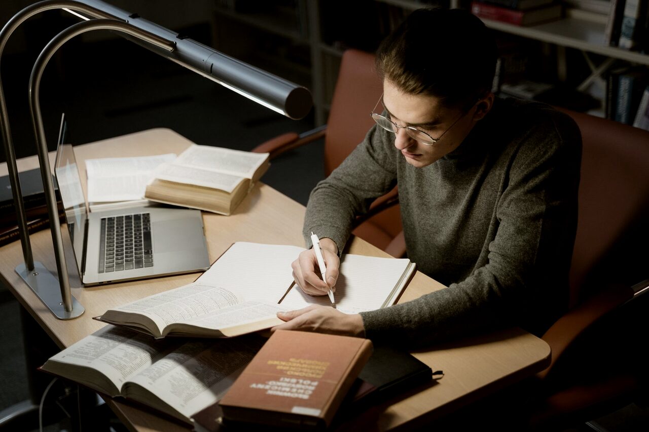 Someone working through a textbook and a laptop by lamplight, taking notes as they go.