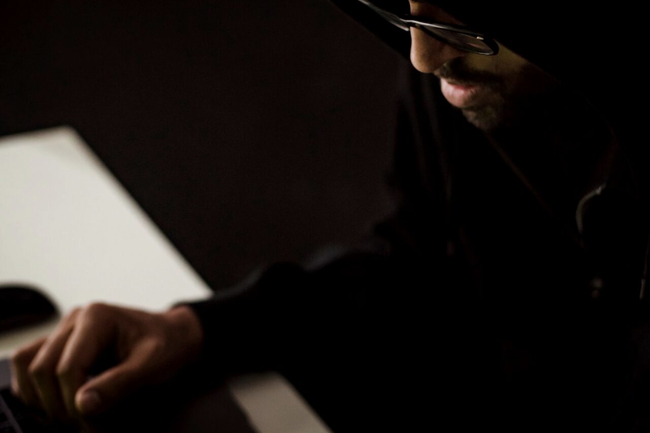 A hooded figure at a laptop in near darkness, face unlit.